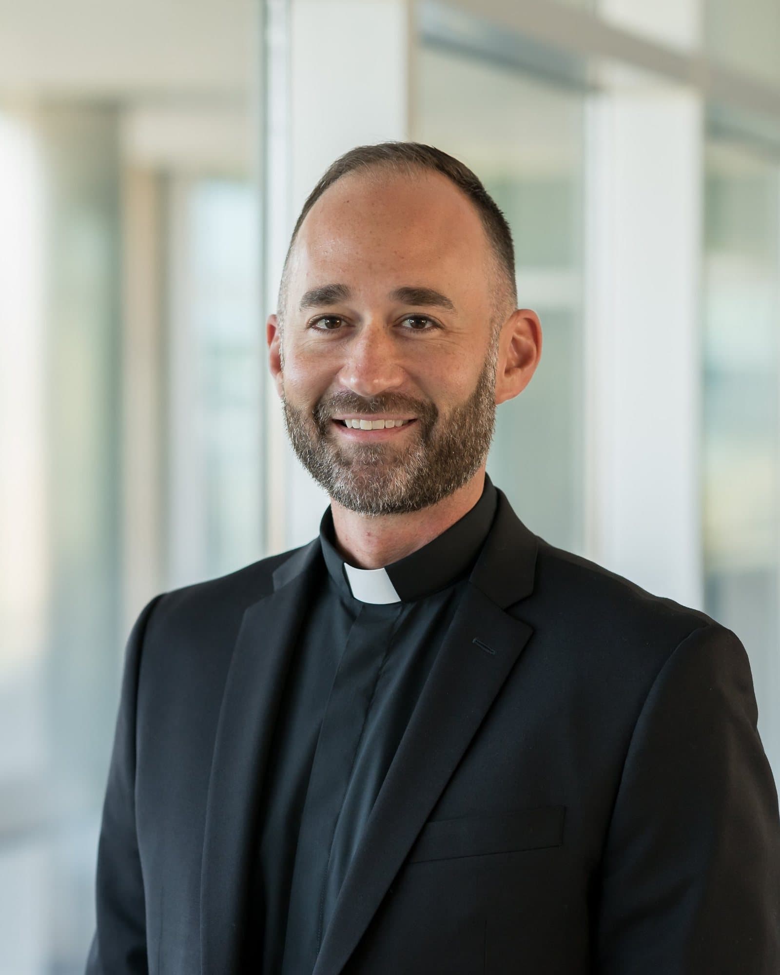 Rev. James Platania, smiling, in a black suit and Roman collar, photographed indoors before a bright glass wall.